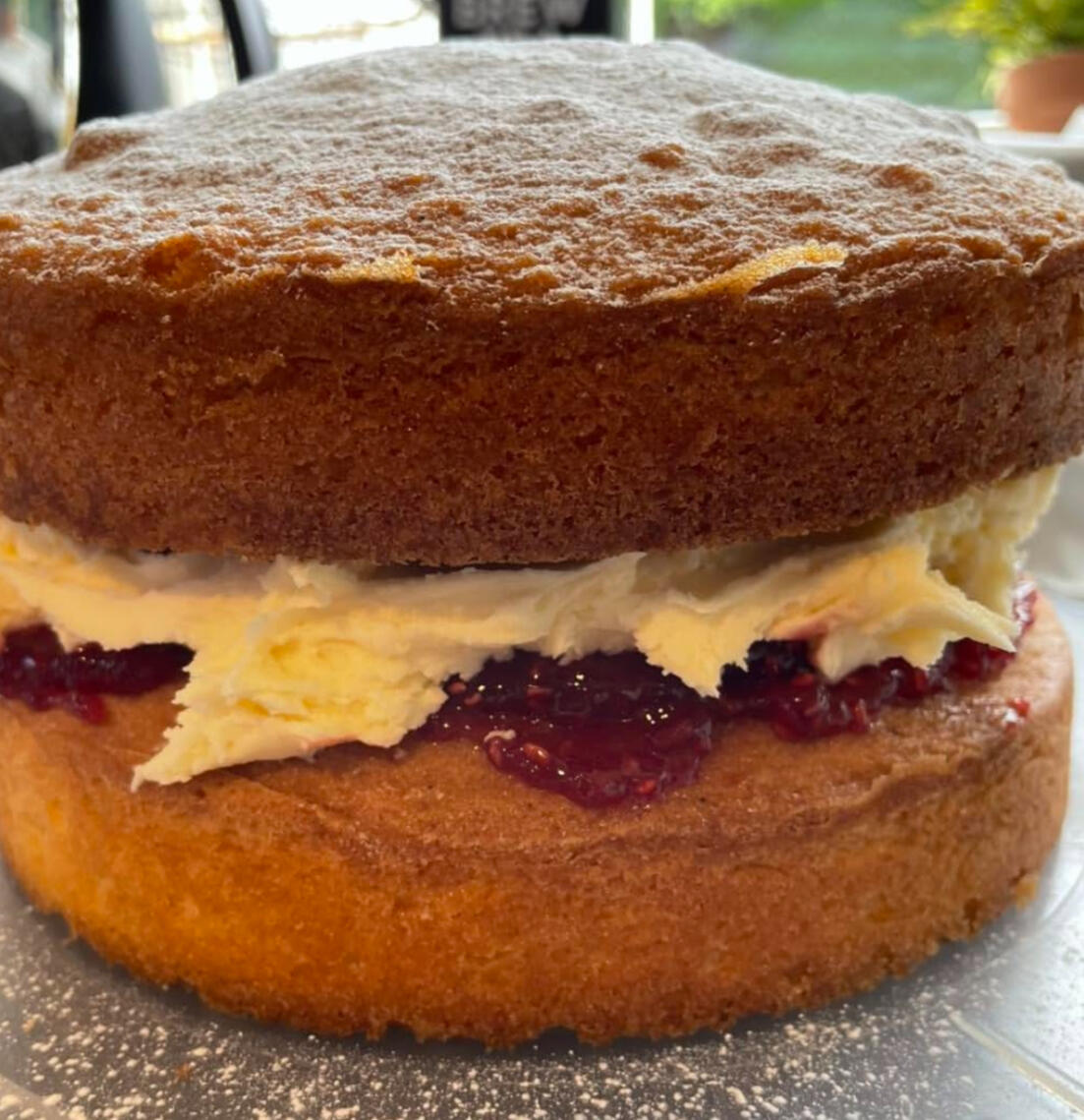 A photo of a victoria sponge on a unit with a sunny window in the background. The cake shows a large filling of buttercream and deluxe raspberry jam